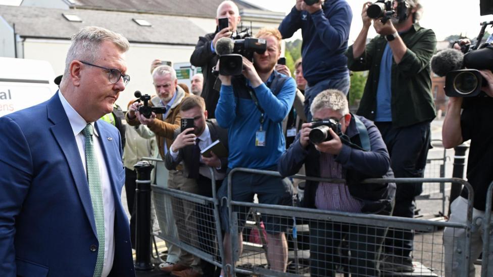 Jeffrey Donaldson, a man with short grey hair and glasses is walking into a courthouse. Behind him are a bank of photographers taking his picture. He is wearing a dark blue suit, a white shirt and green tie.