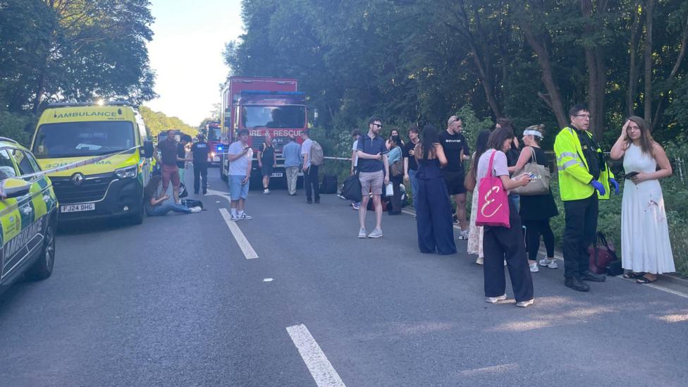 A large group of people standing in a road, with emergency vehicles around them. Most look shocked and some have bandages and blood on them.