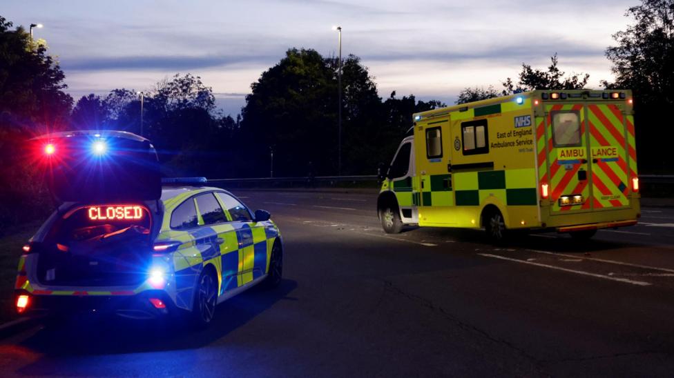 A police car, with a closed sign on it, and an ambulance, on a road, at dusk. Trees are around the road.