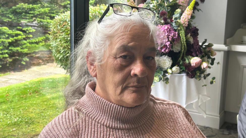 Marie Arbuckle facing the camera. She is wearing a pink jumper, has her hair tied back and is wearing her glasses on her head.
