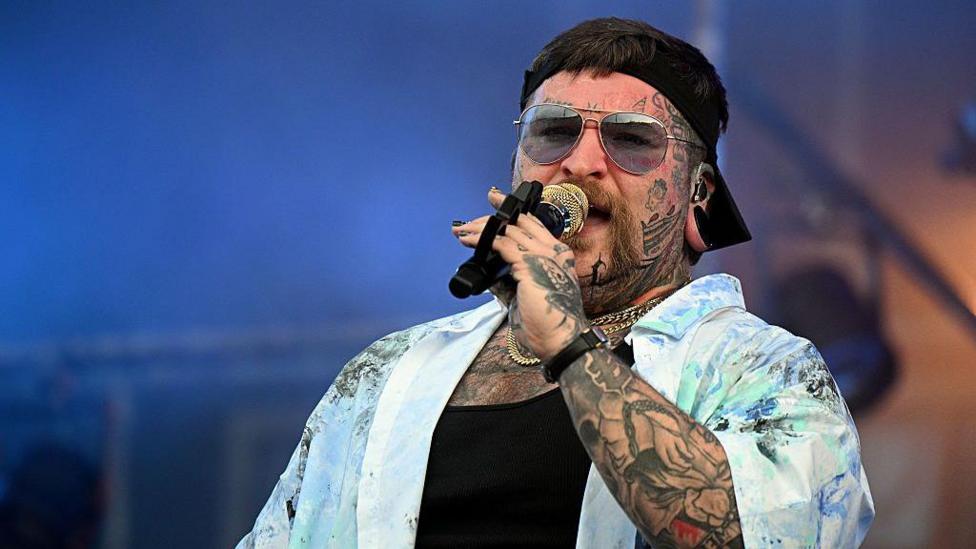 Singer Teddy Swims holding a microphone to his mouth. He is wearing sunglasses, a black cap, a black top and an unbuttoned blue shirt. He has tattoos on his arms and face.