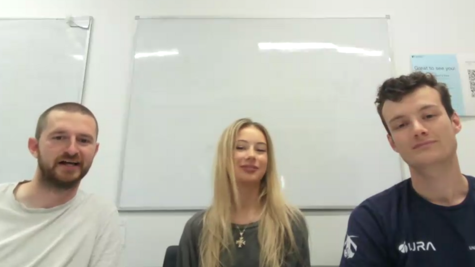 The team members are sitting in a room with a white board behind them. Toby has closely cropped hair, a neatly trimmed beard and he is wearing a white T-shirt. Manon has long blonde hair and she is wearing a dark green coloured top. Seb has dark hair and he is wearing a dark blue T-shirt.