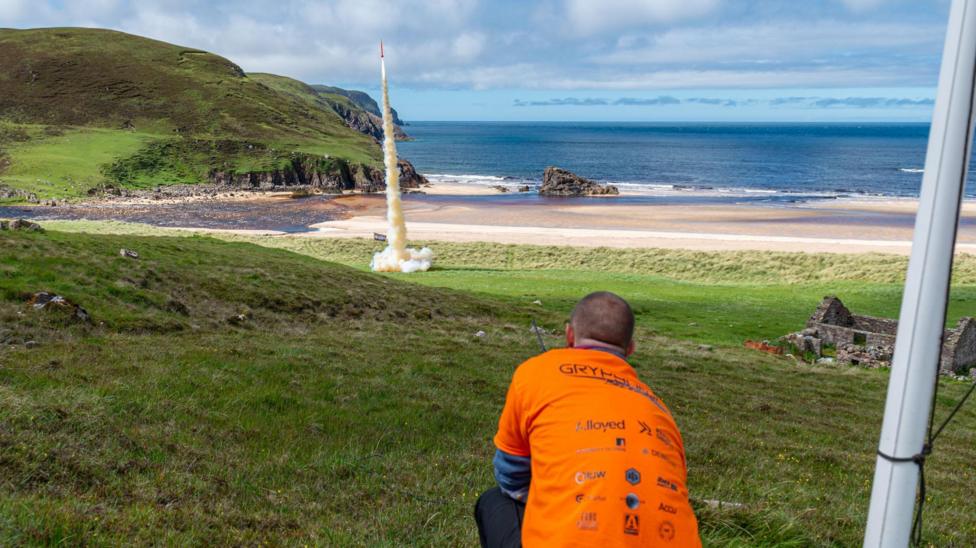 A member of Lura wearing an orange T-shirt crouches down as they watch the orange G2 rocket launch from an area of shoreline at Cape Wrath. The rocket leaves a trail of white smoke.
