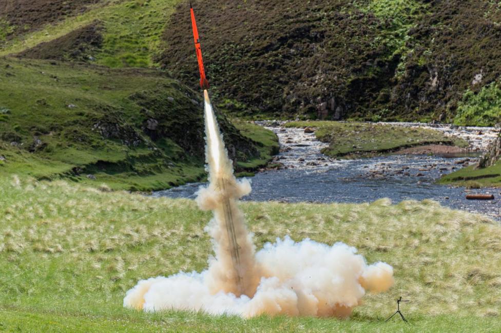 The bright orange rocket leaves a trail of white smoke as it launches from a grassy area.