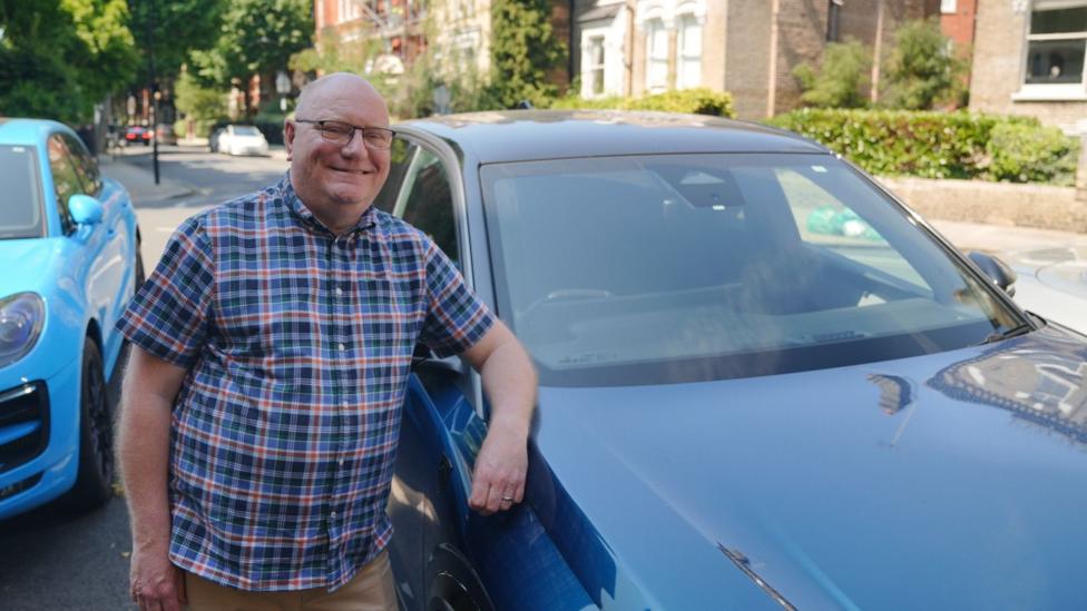 A smiling man in a checked blue and red shirt, and brown trousers leans on his dark blue car parked in the middle of the street. To his right is a lighter blue car and in the background a residential street with other cars driving both ways and front garden hedgerows along the pavement