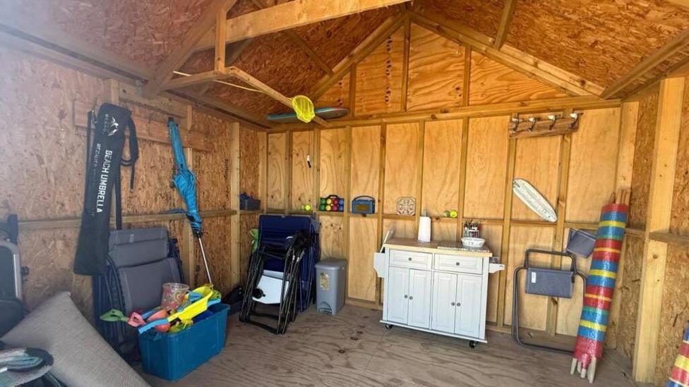 Inside of the wooden beach hut shows a large open-plan space with various different beach items including chairs, wind shield and toys. 