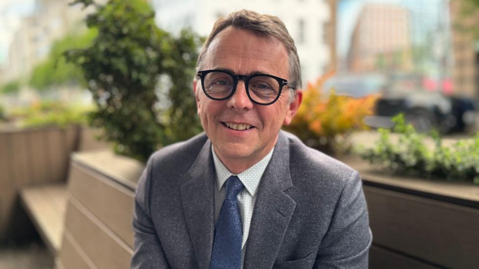 Professor Robbie McDonald. He has short brown hair, wearing glasses, a grey blazer, white shirt and blue tie.