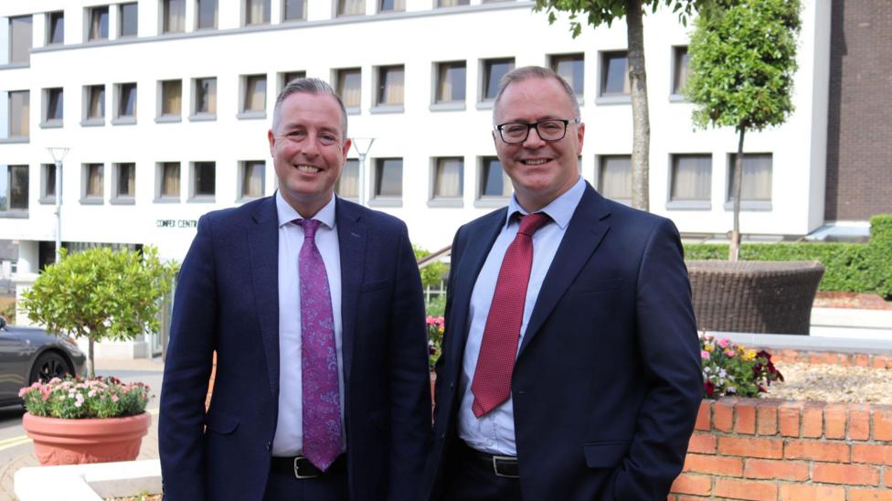 Paul Givan wears a dark suit with a purple tie. Tom Bennett wears a dark suit with a red tie. Both have grey hair and Bennet is wearing glasses. They are standing outside in front of a white building,