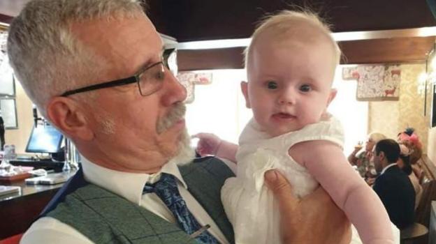 Colin Thwaites is pictured at a function with a grandchild. Colin is wearing glasses and a Harris Tweed waist coat over a white shirt and a dark blue tie.