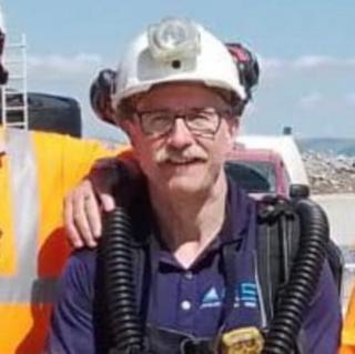 Colin Thwaites wearing a white hard hat with a lamp fitted to the front. He is standing outdoors and it is a sunny day.