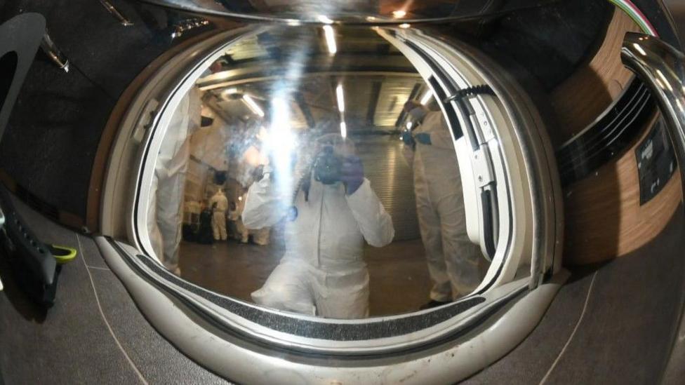 A stainless steel teapot. The reflection shows a man taking a photograph of the teapot.