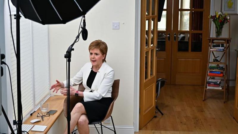 Nicola Sturgeon sitting on a chair next to the window in her house. She is wearing a white jacket and black dress. The library ladder is in the background.