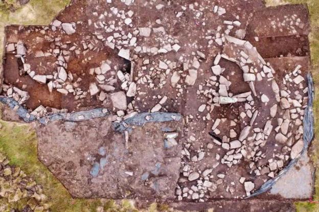 The aerial view of the prehistoric structure at Spurness shows large stones laid out on an area of red soil. Some of the stones have been laid out in such a way they look like walls and chambers. 