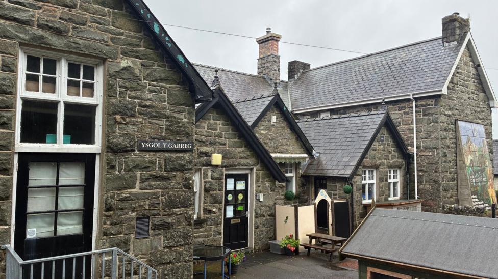 General view of Ysgol Y Garreg, a stone building with black doors and white windows - the building is in the traditional school format of gabled ends and a central section fronting onto a small yard