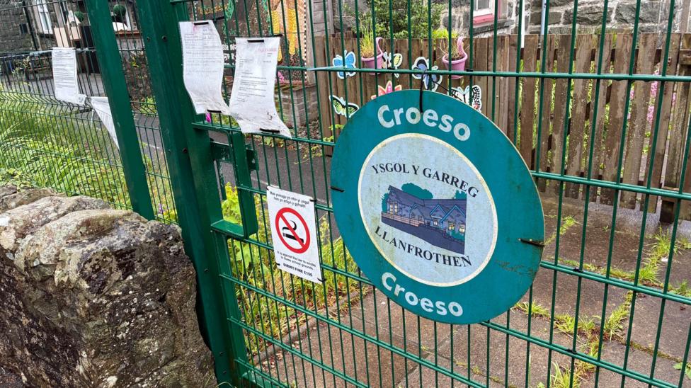 The closed gate of Ysgol Y Garreg, a modern, green wire gate set into a fence of the same type. A modern notice with the school name on - slightly worn - and a no smoking sign are attached to the gate by cable ties
