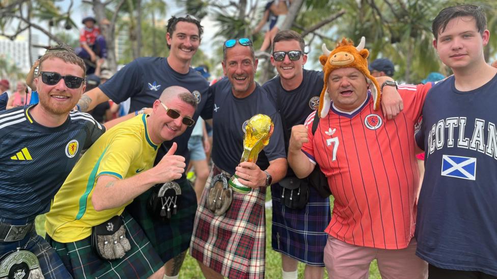 Men in kilts and Scotland tops pose for the camera with their arms around each other