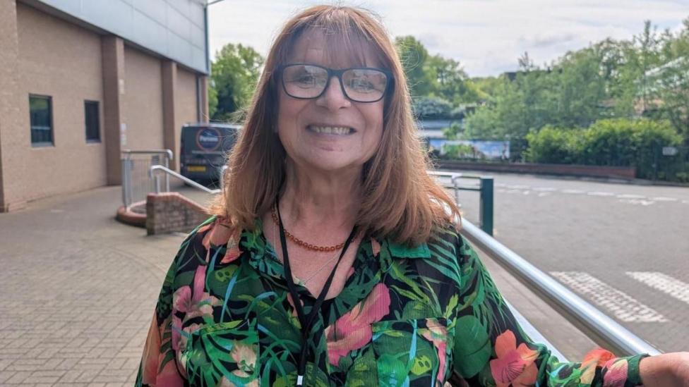 A woman with long brown hair, glasses and a green floral dress smiles as she leans on a hand rail outside a building.