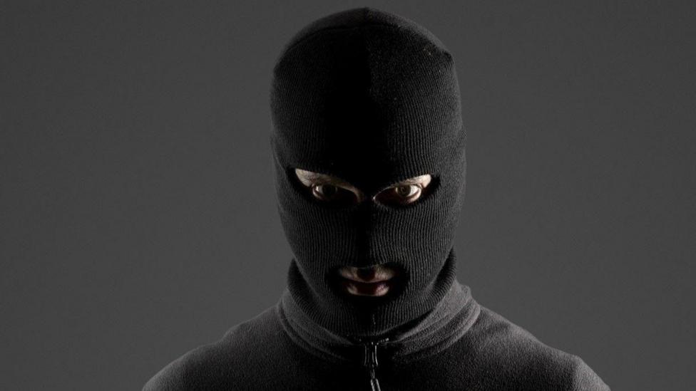 A person stares at the camera while wearing a full-face balaclava and a zipped up, grey jumper. They are standing in front of a dark grey wall.