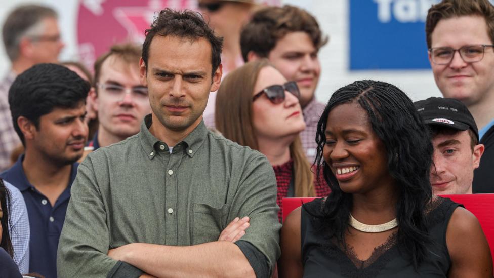 Josh Simons, in a green shirt with a button down collar, folds his arms and looks on as Burnham delivers a speech to supporters. A smiling Miatta Fahnbulleh, in black top, is stood next to him 