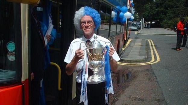 A person stands beside a bus decorated with blue and white balloons, holding a large silver trophy with ribbons.
