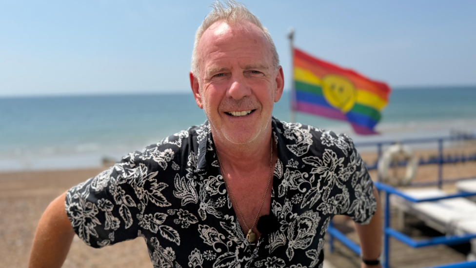 Norman Cook leans against a barrier overlooking Brighton Beach. He is smiling and has one of his trademark patterned shirts on. Behind him we can see the sea and a rainbow flag with a smiley face on it.