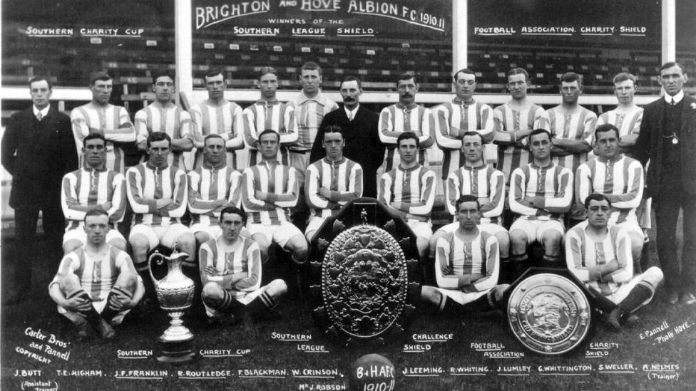 A black and white photo of a football team sitting behind a trophy.