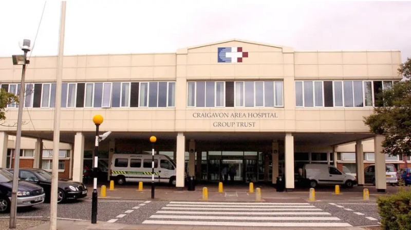 A big off-white hospital facade with a medical crest below the ridge. Parked out the front are ambulances and there is a zebra crossing in foreground, 