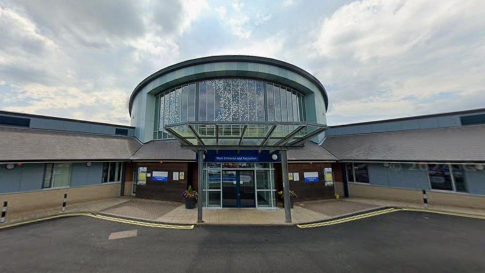 Lanchester Road Hospital's main entrance. It it a single-storey building with a large round atrium with huge windows on the roof behind the front door. Two wings fan out at 45-degree angles from the central entrance which has automatic sliding doors.