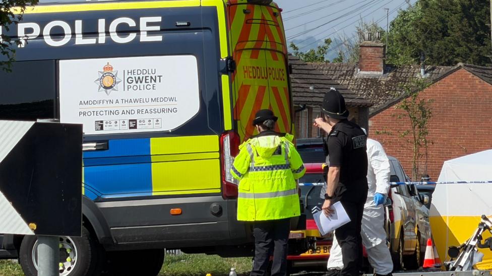 Emergency services staff and vehicles on Aberthaw Road. There is a police van with one officer with her back to the camera in a fluorescent coat, and another in a police hat, black t-shirt and black vest. A police cordon and white tent can be seen in the background. 