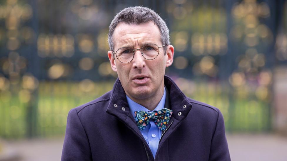 Muir is outside. He's talking. He wears glasses. He has a navy coat on with a blue shirt and floral bow tie.