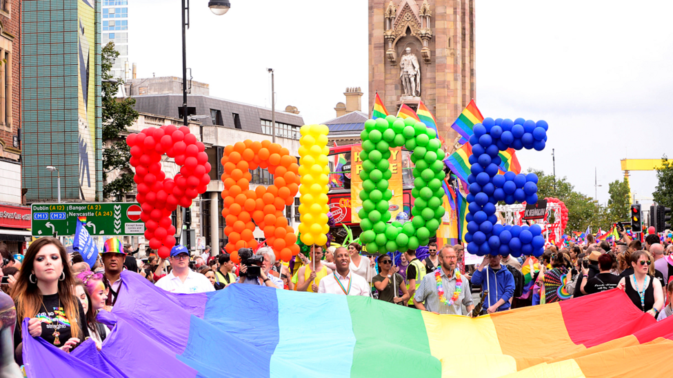 A large rainbow flag is being held by people taking part in a parade. The Albert clock is in the background. Pride balloons are being held up.
