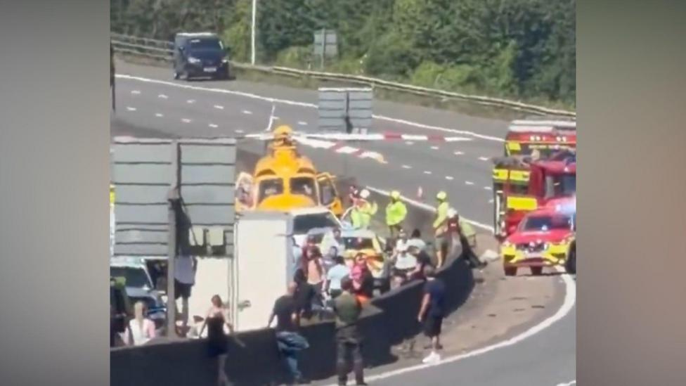 Emergency services and motorists spending on a motorway. There is a yellow helicopter nearby and two fire brigade vehicles.