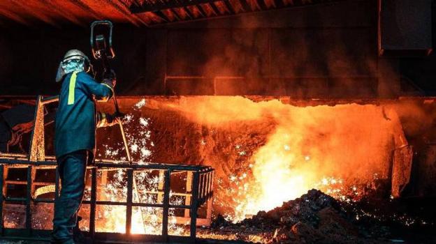A worker at the blast furnace. They stand on a platform, wear a helmet and protective clothing, and hold a long bar into a pit below. Sparks and orange smoke can be seen the background. 