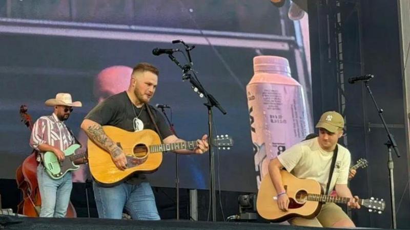 The photo is of Paul performing with Zach Bryan. They are standing on a stage and are both holding brown acoustic guitars. 