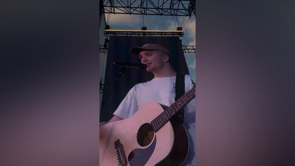 The image is a photo taken of a screen at a concert.It shows Paul with a brown acoustic guitar. 