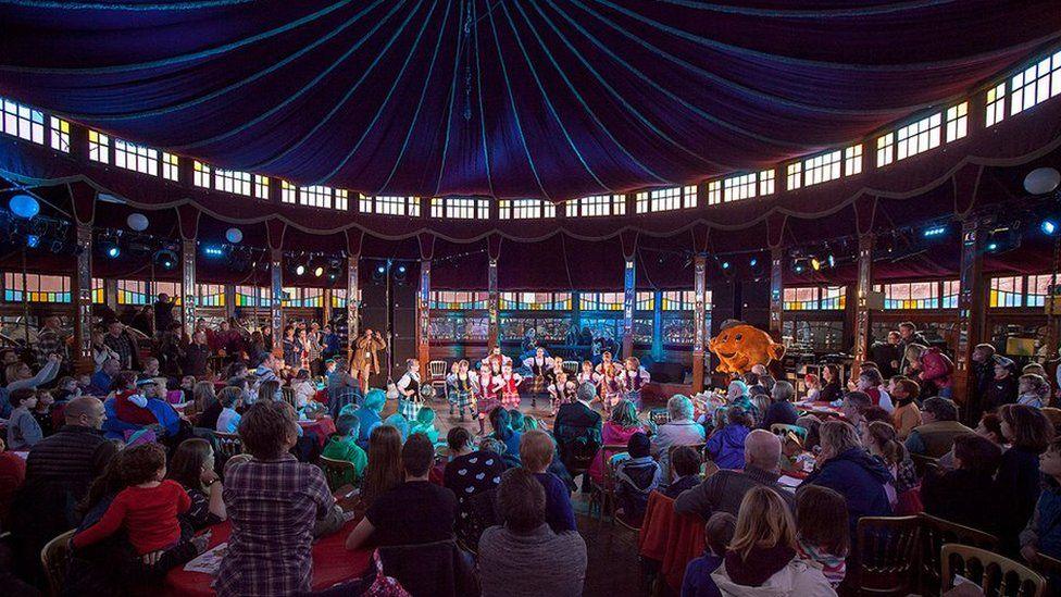 A view of some people doing Scottish country dancing in a large tent-like building with a big crowd looking on