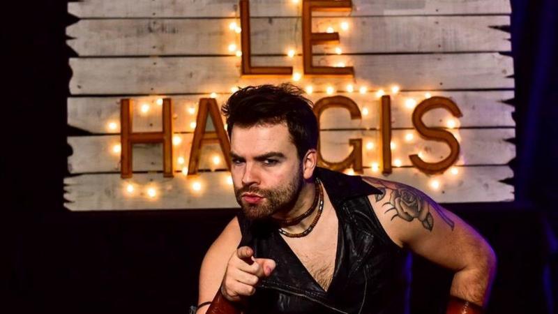 A man with dark hair and a beard and a rose tattoed on his shoulder points at the camera with a sign saying Le Haggis lit up behind him