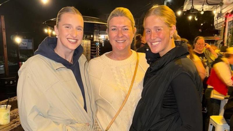 Three women looking at the camera and smiling. The pic is taken outdoors at night.