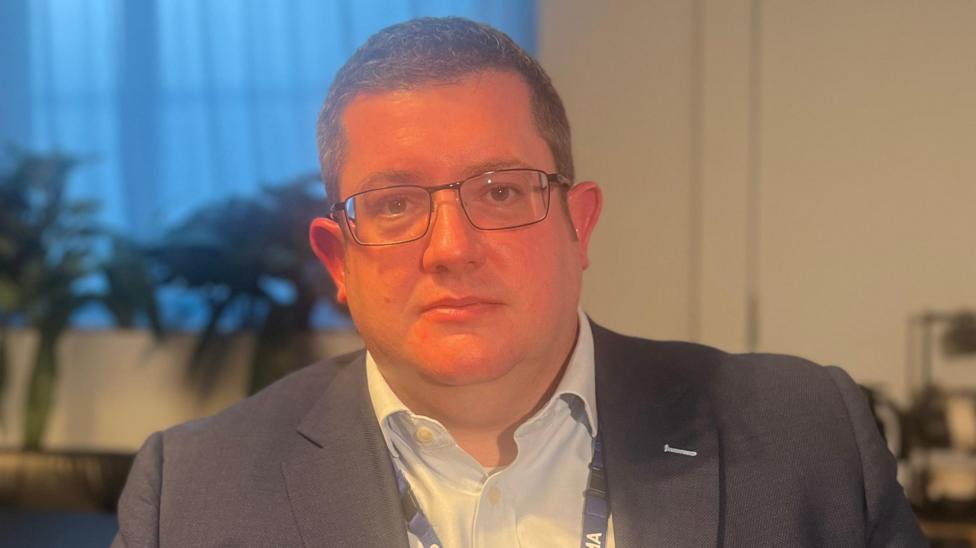 A man with dark hair and glasses, he is wearing a navy blazer and white shirt.