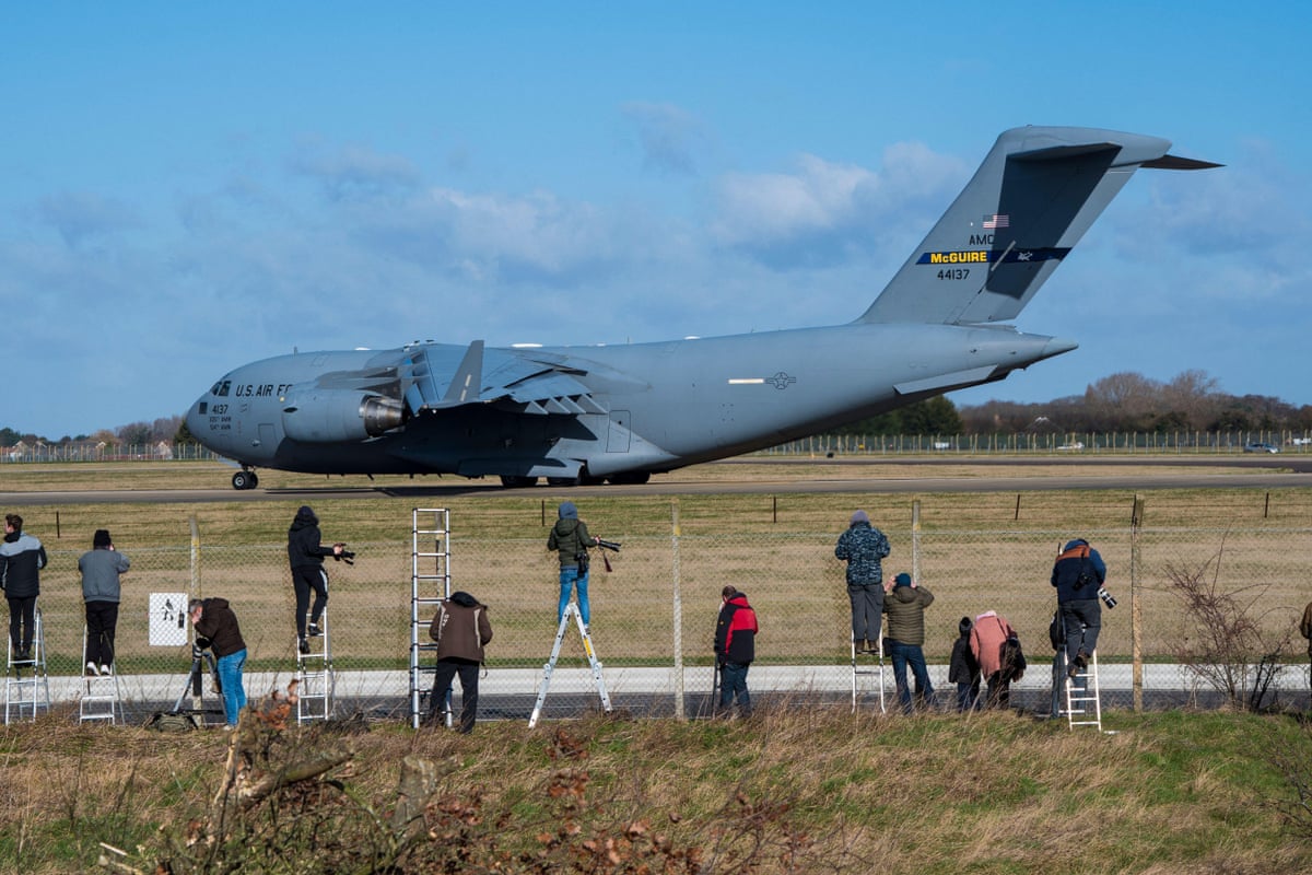 People on ladders and standing at a fence photographing a large military transport aircraft on an airfield