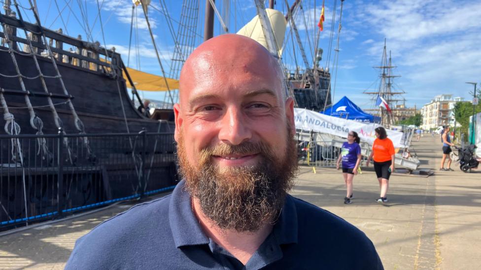 A smiling Lupo Bathke stands on a quay in front a replica Spanish galeon. He is smiling and wearing a blue polo shirt. He is bald, two women can be seen in the background
