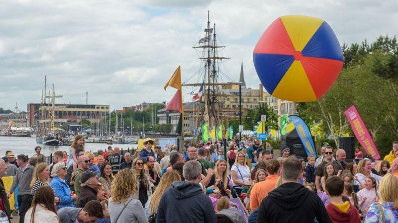 a large crowd of smiling people gather on the quay in Derry enjoying the martime festival's activities. A large, inflatable ball which is yellow, blue and red is floating in the air while ships and the River Foyle can be seen in the background