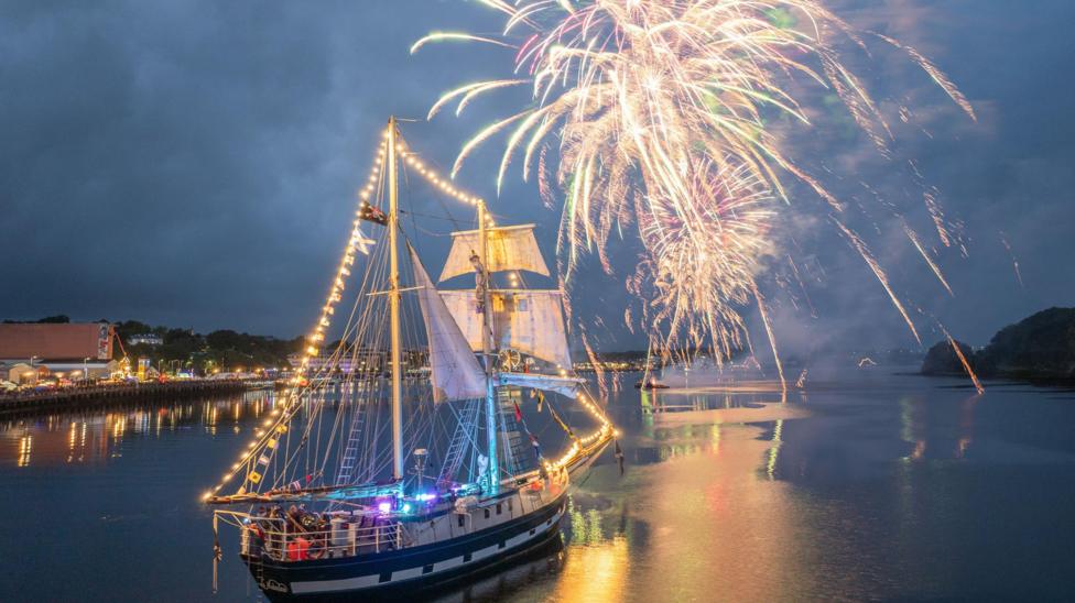 fireworks go off above the river Foyle , a tall ship is in the foreground. It is dark and the ship is lit up 