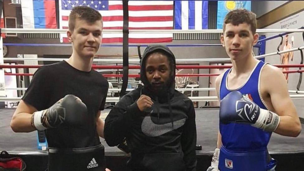 Aaron and Stephen McKenna stand with rapper Kendrick Lamar inside a Los Angeles boxing gym, with a boxing ring and national flags visible behind them.