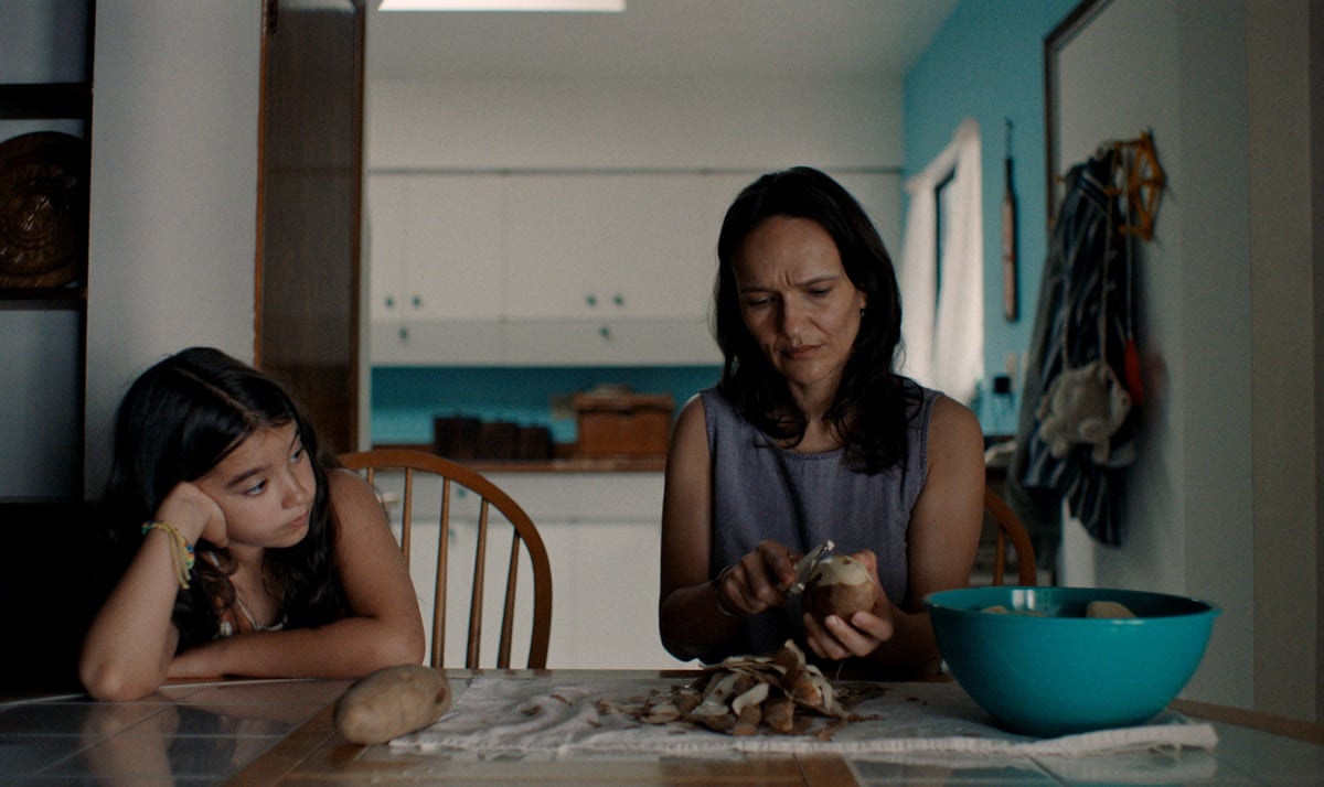 A young girl sits at the kitchen table while her mother peels potatoes in Blue Heron