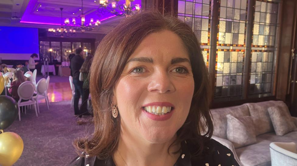 Marion McMahon is standing in an events space with different tables set out and a blue projector screen. A sofa and chairs are visible to her right. She has brown hair, earrings and a black dress with small white hearts on it