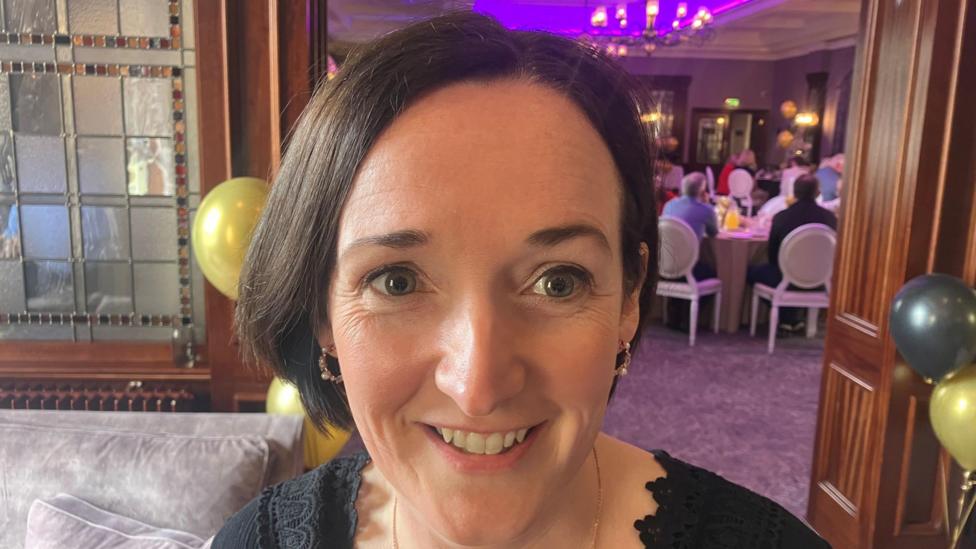 Edel McGeeney is standing in an events space with different tables set out and a blue projector screen. A sofa and chairs are visible to her left. She has short dark hair and a black dress.