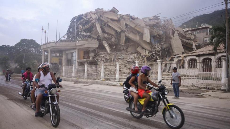 People ride past a collapsed building after an earthquake, in La Guaira