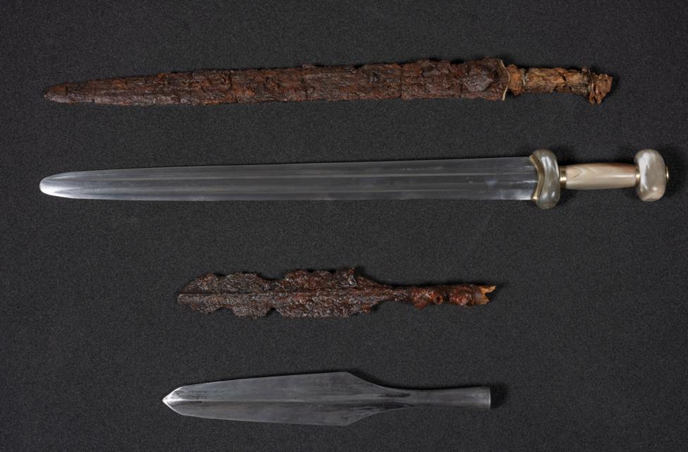 An Iron Age spearhead and sword, both heavily rusted with age. Modern replicas are displayed beside them