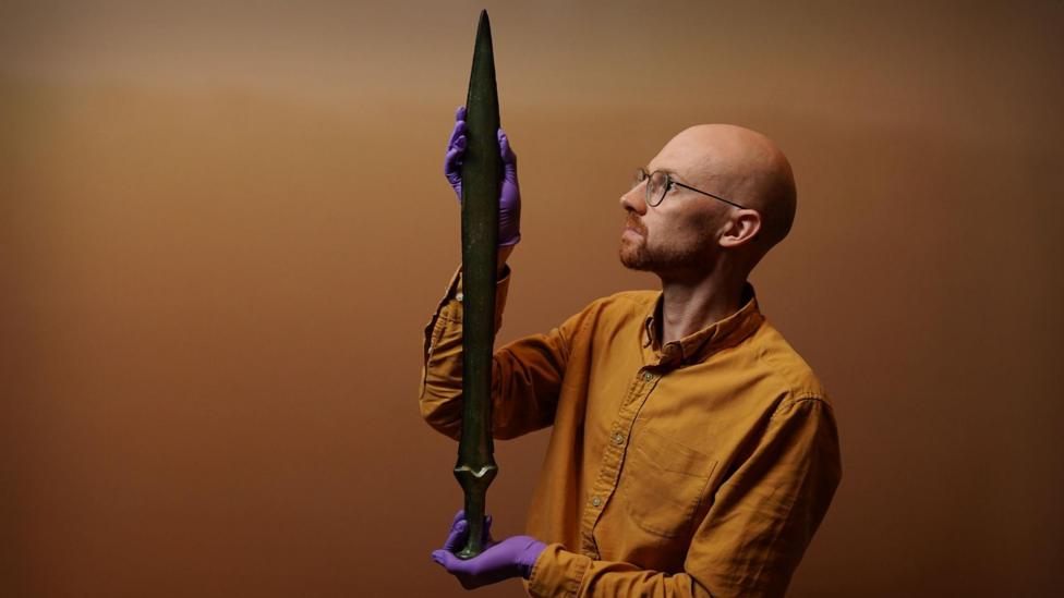 Dr Matthew Knight is standing in a room holding the sword. Dr Knight has a bald head, a neatly trimmed beard and is wearing a mustard coloured shirt, and glasses. He is wearing purple, rubber gloves.